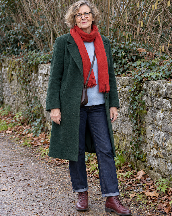 Femme aux cheveux gris en manteau vert, écharpe rouge, pull bleu clair et jean brut