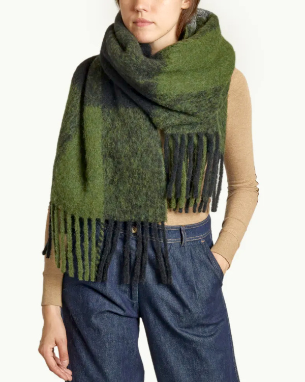 Femme portant une grande écharpe duveteuse verte et gris foncé à franges, avec un haut beige et un jean